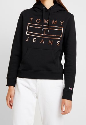 Schwarzer Hoodie mit metallic roségoldenem „TOMMY JEANS“-Logo und Fronttasche, getragen mit weißer Hose.
