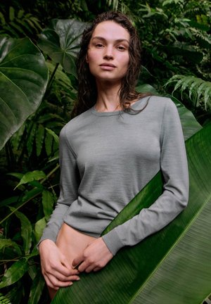 Kvinde med vådt hår iført en lysegrå langærmet top, stående blandt store grønne tropiske blade, med hænderne hvilende nær hendes talje.