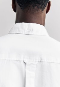 Rückansicht einer Person mit kurzen lockigen Haaren, die ein weißes Hemd mit einem Knopf am hinteren Kragen trägt.