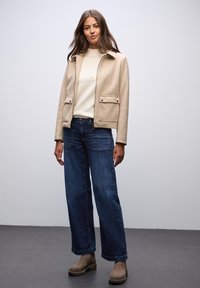 Beige ulljacka med krage och två framfickor, krämfärgad tröja, mörkblå vida jeans och bruna ankelstövlar. Slät textur.