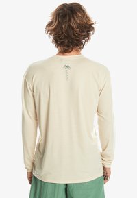 Langärmliges, cremefarbenes T-Shirt aus weichem Stoff. Mit einem kleinen grünen Palmenbaumdesign auf dem Rücken. Figurbetonter Schnitt.