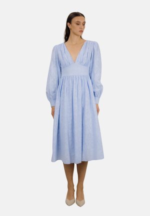 Kvinne iført en lyseblå midikjole med dyp V-utringning, lange puffete ermer, innsvinget midje og beige spisse hæler.