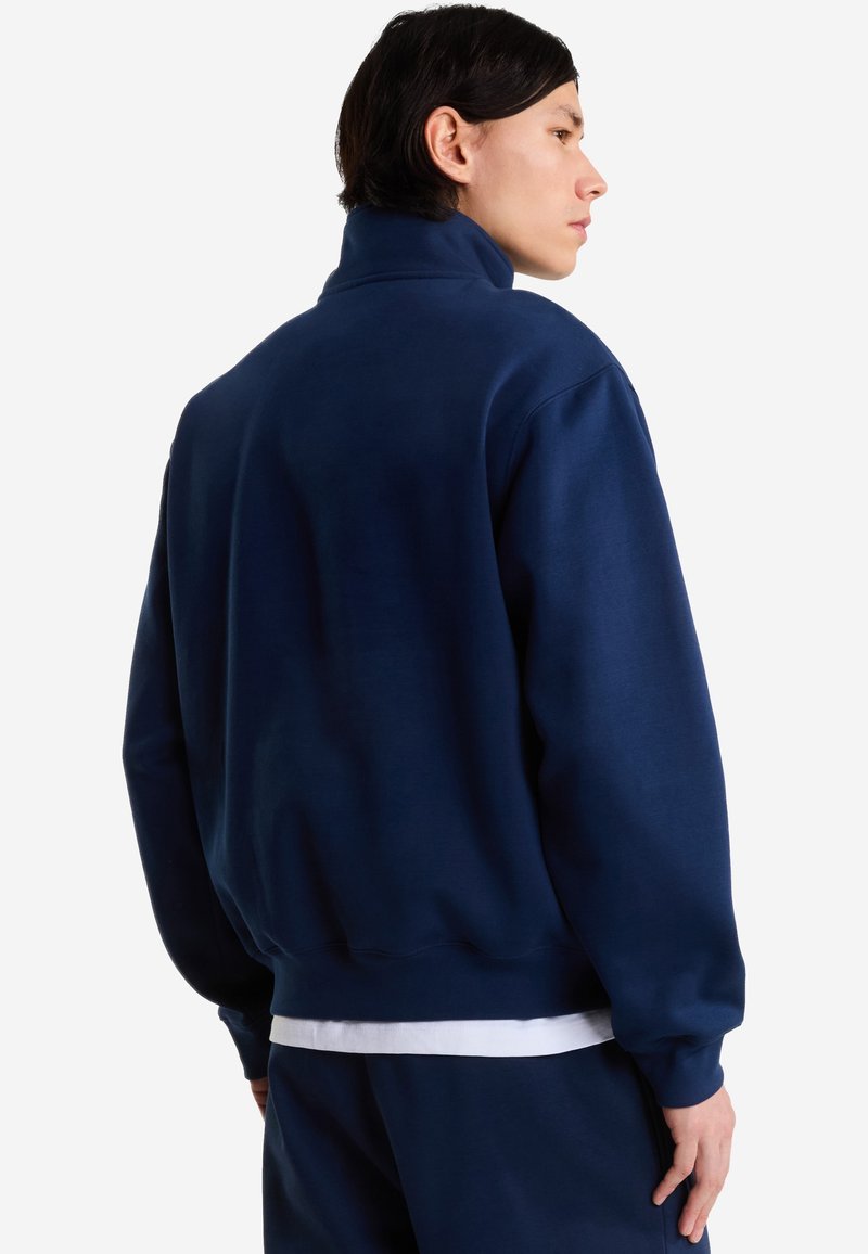 Hombre de perfil con una sudadera azul marino de cuello alto y pantalones a juego sobre una camisa blanca.