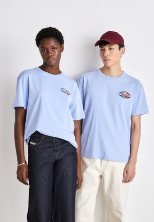 Camisetas de algodón de color azul claro con bordado de logo en el pecho, usadas con jeans oscuros y pantalones de color claro, ambos modelos de pie uno al lado del otro.