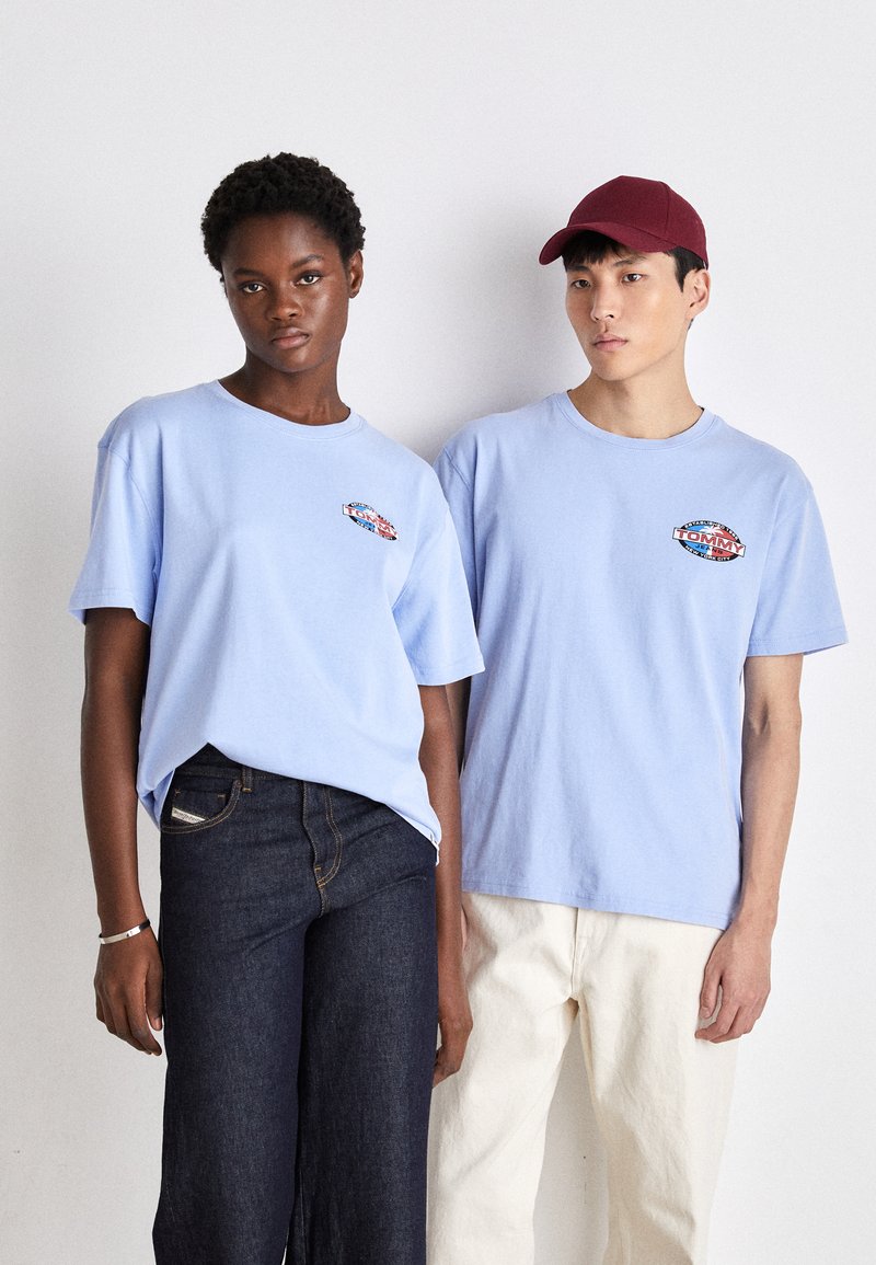 Camisetas de algodón de color azul claro con bordado de logo en el pecho, usadas con jeans oscuros y pantalones de color claro, ambos modelos de pie uno al lado del otro.