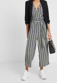 Tuta a righe verde e blu navy con design avvolgente, gambe larghe e cintura legata, abbinata a un blazer nero e sneakers bianche.