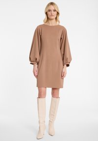 Robe longue marron à manches ballon, décolleté rond, faite dans un tissu doux ; associée à des bottes beiges claires jusqu'aux genoux.