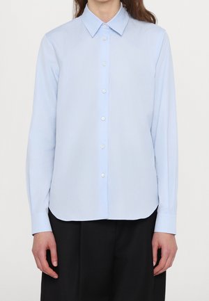 Kobieta ubrana w jasnoniebieską koszulę na guziki z długim rękawem i zaostrzonym kołnierzykiem, w połączeniu z czarnymi spodniami, na jednolitym tle.