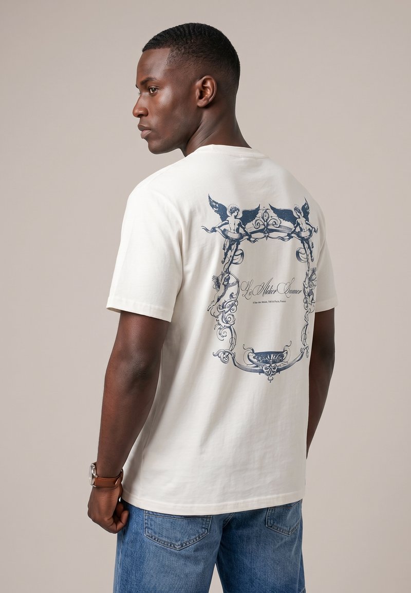 Homme aux cheveux courts portant un t-shirt blanc avec un chérubin bleu et un cadre orné imprimé dans le dos, associé à un jean bleu et une montre-bracelet marron.