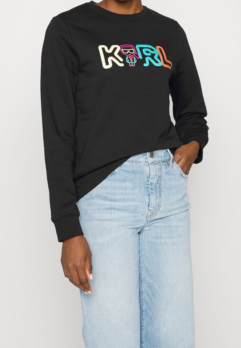 Personne portant un t-shirt noir à manches longues avec un texte coloré "KARL" et un petit dessin de personnage, associé à un jean bleu clair.