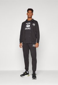 Zwarte trainingspak met een ritssluiting jack en bijpassende broek. Bevat witte logodetails op het jack en de borst. Gedeeltelijk gedragen met zwarte sneakers.