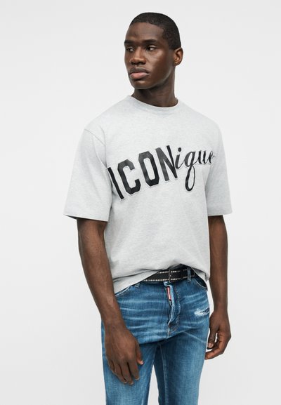 Νέος άντρας με σκούρο χρώμα δέρματος, που φορά ανοιχτό γκρι μπλουζάκι με το κείμενο "ICONique" και μπλε τζιν με φθορές, κοιτάει προς τα αριστερά του.