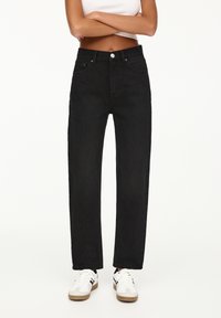 Jean en denim noir avec une coupe droite, cinq poches et une fermeture à bouton. Porté avec des baskets blanches, présentant des accents beige.