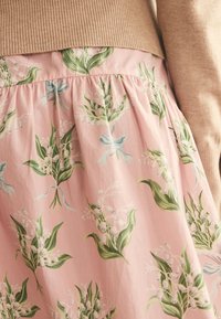 Fustă roz cu model floral alb și frunze verzi, purtată sub un pulover bej cu mâneci cu dungi pe trunchiul unei persoane.