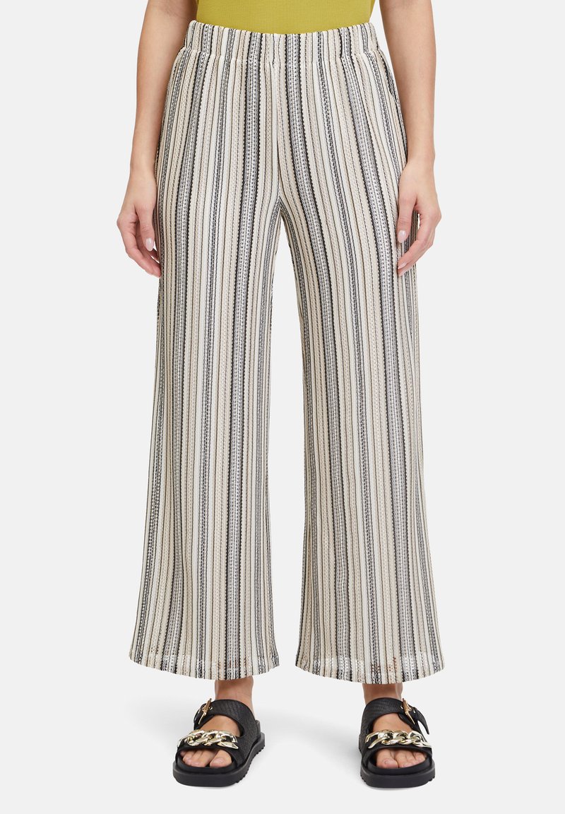 Wijdvallende broek in crème met zwarte verticale strepen. Elastische tailleband. De stof heeft een subtiele textuur. Samen gedragen met zwarte sandalen met een gouden ketting.