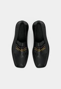 Mocassins noirs avec une finition en cuir lisse, des détails à franges et un accent de chaîne dorée sur le cou-de-pied. Conception à bout carré.