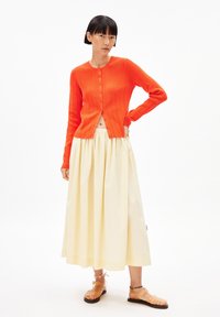 Helles orange gestreiftes Strickjäckchen mit Rundhalsausschnitt und Knöpfen vorne, kombiniert mit einem fließenden cremefarbenen Rock und transparenten Sandalen.