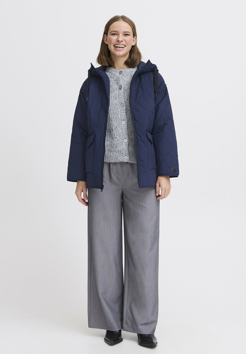 Giacca trapuntata blu navy con cappuccio, maglione grigio a trecce e pantaloni grigi a gamba larga. Il modello è in piedi con le mani ai fianchi, stivali neri visibili.
