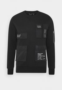 Černá mikina s několika strukturovanými kapsami, každá s bílým tištěným textem. Má kulatý výstřih a kapsu na zip na rukávu.