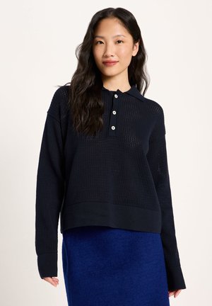 Young woman with long black hair wearing a dark navy knit sweater with buttons and a bright blue skirt against a light background.