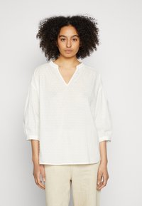 Blusa branca, solta, com decote em V, mangas três quartos, padrão texturizado e um design simples; combinada com calças bege claras.
