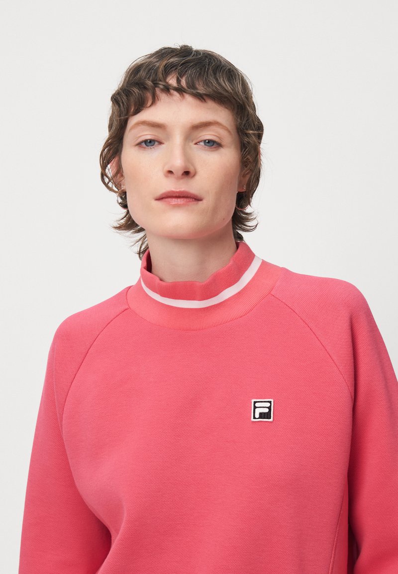 Felpa rosa con colletto alto, tessuto strutturato, maniche raglan e un'etichetta con logo nera sul petto. Presenta strisce bianche decorative.