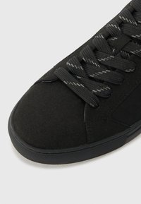 Crne suede tenisice s teksturiranim završetkom, sivi vezice, zaobljeni nos i gumeni potplat. Minimalistički dizajn i elegantan profil.