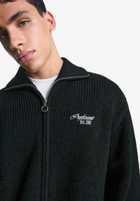 Black knitted zip-up sweater with a high collar, featuring textured fabric and white embroidered logo "Argotient Est. 2001" on the chest.
