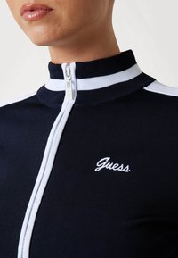 Morska zimska jakna na patent, s bijelim prugama na ovratniku i ramenima, koja sadrži izvezeni logotip "Guess" u bijeloj boji, od teksturiranog materijala.