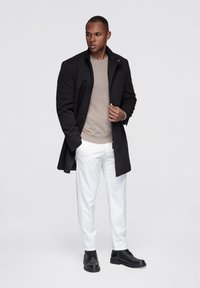 Zwarte wollen jas over een beige trui, gecombineerd met witte broek en zwarte veterlaarzen. Soepele texturen en strakke lijnen zijn duidelijk aanwezig.