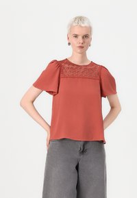 Top corto color ruggine con dettagli in pizzo attorno al collo, realizzato in tessuto morbido. Presenta una vestibilità ampia e un orlo arrotondato.