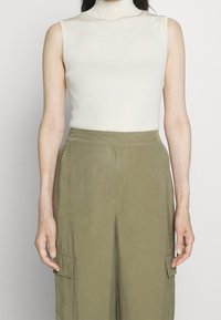 Top senza maniche a collo alto a coste crema abbinato a pantaloni larghi verde oliva con tasche laterali e una trama di tessuto liscia.