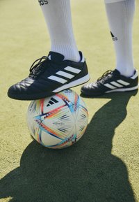 Svarta träningsskor med vita ränder på sidan, placerade på en färgglad fotboll, mot en grön gräsmatta som bakgrund.