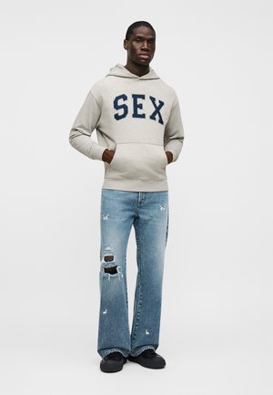 Szürke gyapjú kapucnis felső tengerészkék betűkkel "SEX," világoskék szakadt bőszárú farmerrel és fekete cipővel párosítva. A modell fehér háttér előtt áll.