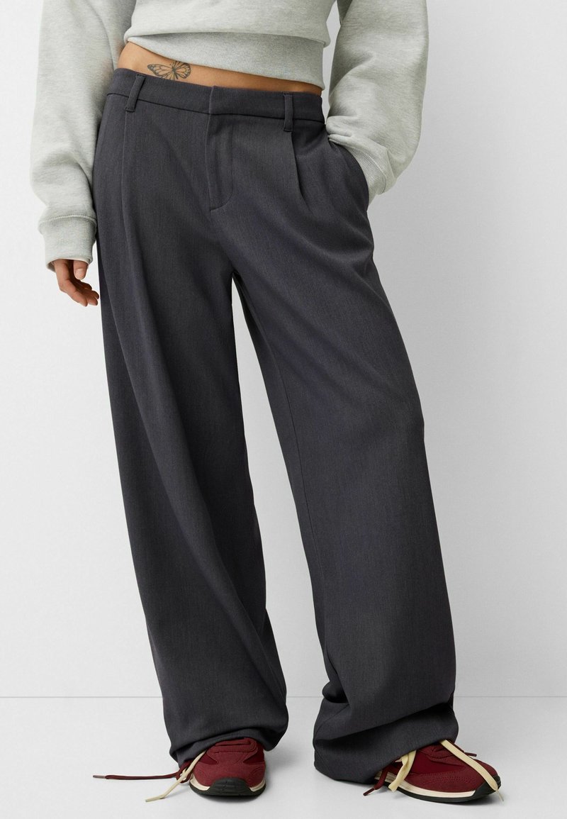 Pantalons gris foncé à jambes larges avec une coupe ajustée, des plis à l'avant, et une texture lisse. Associés à des chaussures de sport rouges avec des accents légers.
