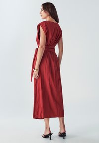 Červené, bezrukávové maxi šaty s páskem v pase, s drapérií látky a jemnou texturou. Mají otevřený zadní díl a délku ke kotníkům.