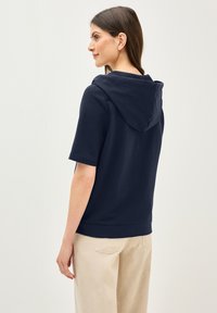 Sweat-shirt à capuche bleu marine à manches courtes et coupe décontractée, présenté de dos. Tissu doux avec une texture lisse. Pantalons de couleur claire.