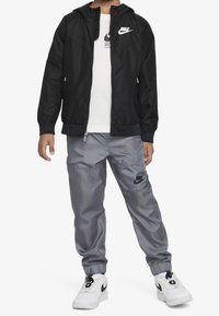 Barn klädd i en svart Nike-jacka, vit Nike-t-shirt, grå Nike-joggingbyxor och vita Nike-sneakers, stående mot en vit bakgrund.