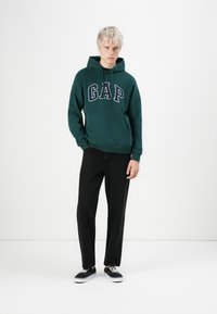 Green GAP hoodie with front kangaroo pocket, bold logo in navy, paired with black pants and black sneakers, standing on a white background.