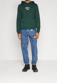 Grön huvtröja med framficka och texten "DENVER". Blå jeans med lappar och märken. Svarta sneakers med strukturerade sulor.