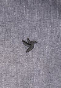Grå stof med fin tekstur, der har en sort kolibri broderi, placeret off-center. Syningerne har klare, veldefinerede kanter.