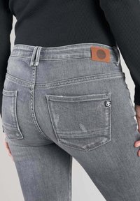 Jean en denim gris avec une légère décoloration, deux poches arrière et une patch en cuir marron à la taille. Doté d'un design ajusté et de détails cousus.