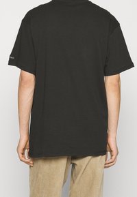 T-shirt noir à manches courtes en coton, avec une coupe décontractée et un col rond. Branding minimal sur la manche gauche.