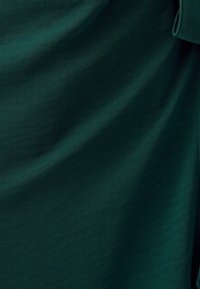 Tessuto verde scuro con una trama liscia, che mostra pieghe sottili e un accento legato su un lato, enfatizzando il suo design fluido.