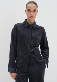 Giacca in velluto a coste grigio scuro con chiusura a bottoni, colletto a punta, due tasche frontali e tessuto dalla texture particolare. Presenta maniche lunghe e una vestibilità rilassata.