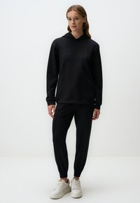 Zwarte hoodie met een losse pasvorm en een voorkantzak, gecombineerd met zwarte getailleerde joggers met elastische boorden. Gecombineerd met witte sneakers.