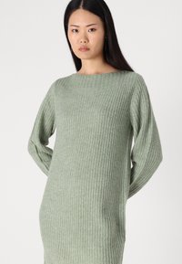 Pull en tricot de couleur vert clair avec un col large, des manches longues et un motif de côtes texturé. Coupe décontractée, matière douce.