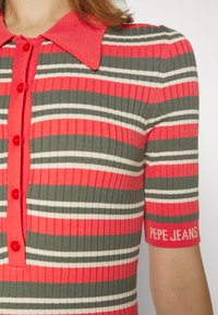 Blouse tricotée rayée aux couleurs rouge, verte et blanche, manches courtes, col pointu avec boutons sur le devant. Logo sur la manche.