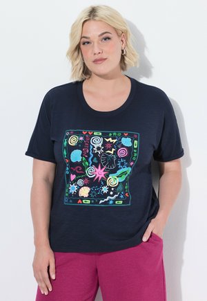Kvinde med kort blondt hår iført en mørkeblå t-shirt med farverige ferietema-doodles og magenta bukser, stående mod en hvid baggrund.