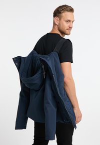 Giacca da esterno blu navy con cappuccio, appoggiata sulla spalla. Presenta una chiusura con zip e bottoni, con una texture di tessuto liscia.